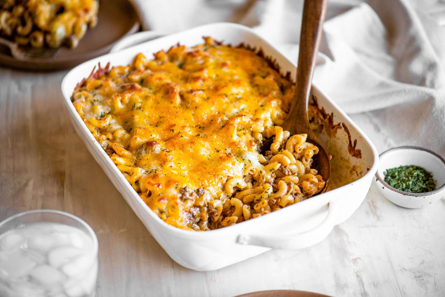Casserole Dish of Cheeseburger Casserole with a Serving Already Served Using a Spoon at a Table Setting (a Plate with a Serving of Casserole, Bowl of Dry Parsley, a Table Napkin, and a Glass of Ice Water)