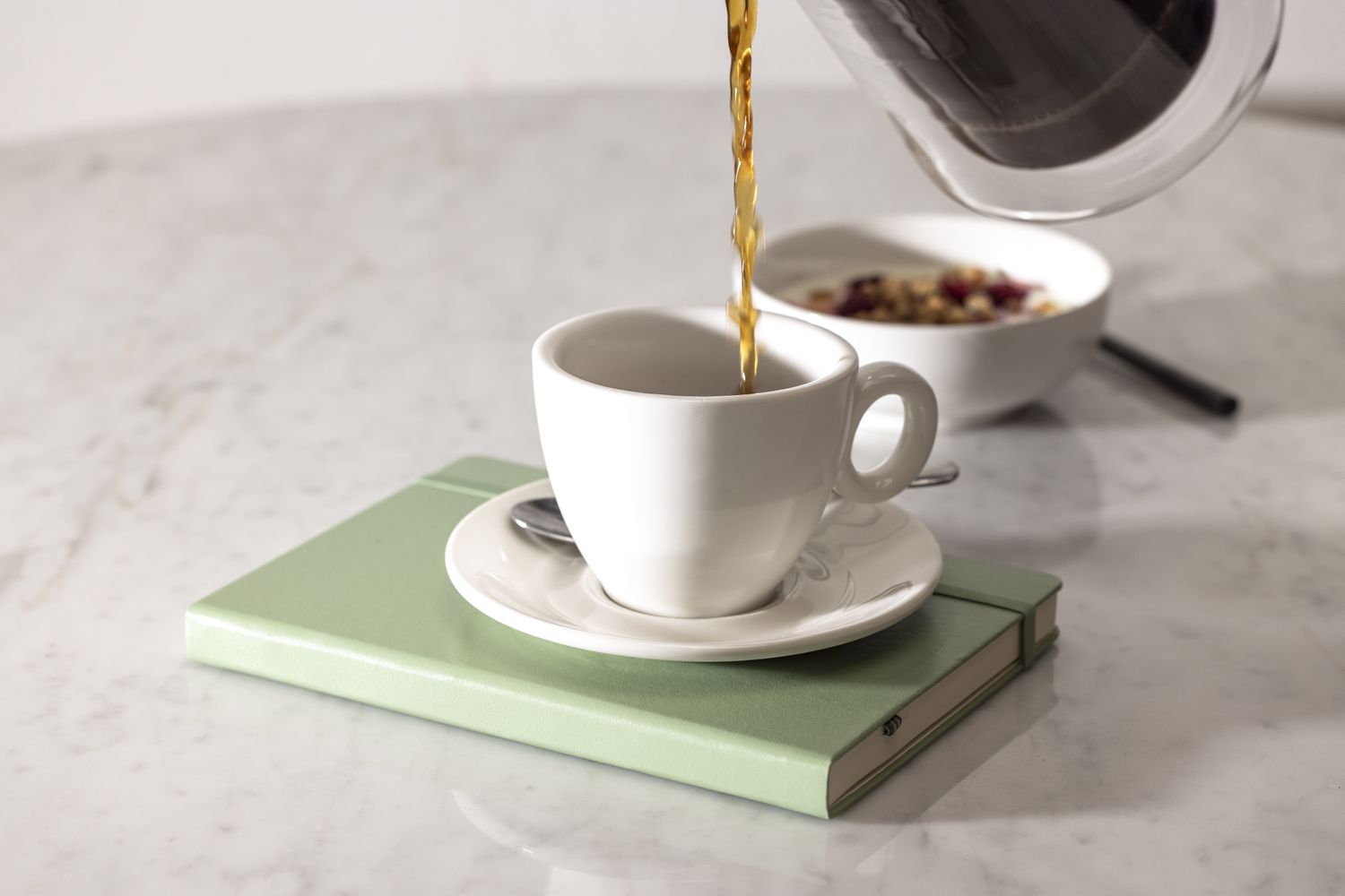 Coffee being poured into a white coffee mug that is on top of a green colored book all on a marble tabletop