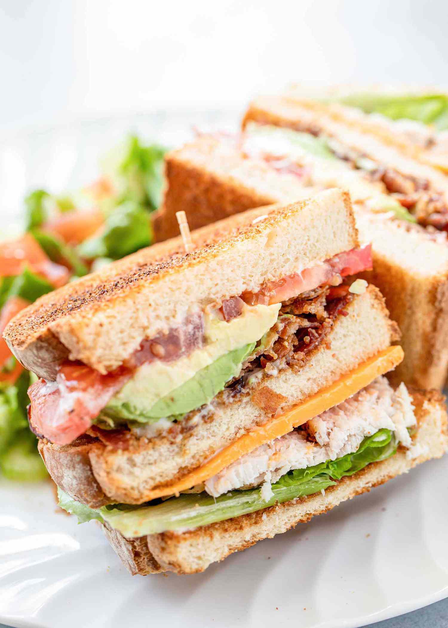 Side view of turkey club with garlic mayo on a plate. The bread is toasted and the mayo, avocado, bacon, tomato, lettuce and turkey are visible in the sandwich. A second half of the sandwich is behind the first.