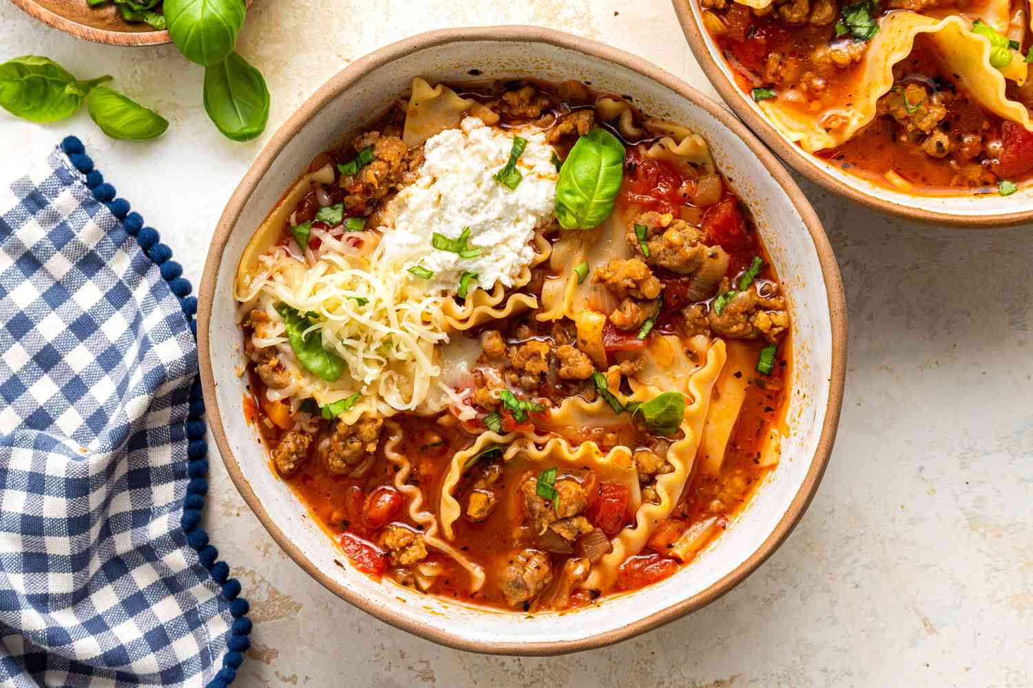 Lasagna Soup in a Bowl With Noodles, Sausage, Ricotta, and Fresh Basil, and in the Surroundings, Another Bowl With All the Toppings, a Saucer With Basil Leaves, and a Blue and White Checkered Kitchen Towel 