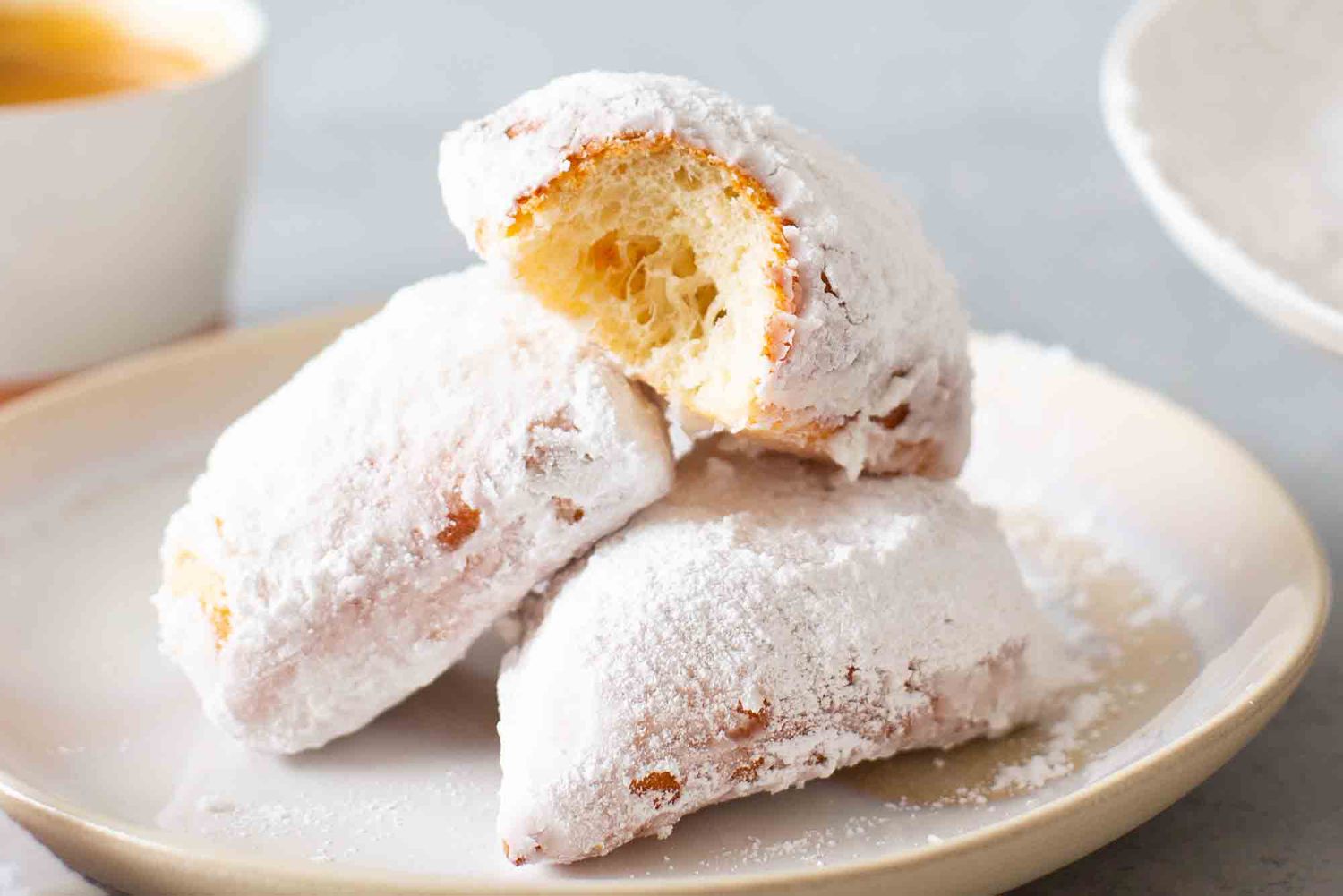 Three Cafe Du Monde Beignets on a cream plate with a cup of espresso to the left. A partial view of a cake platter is on the right.