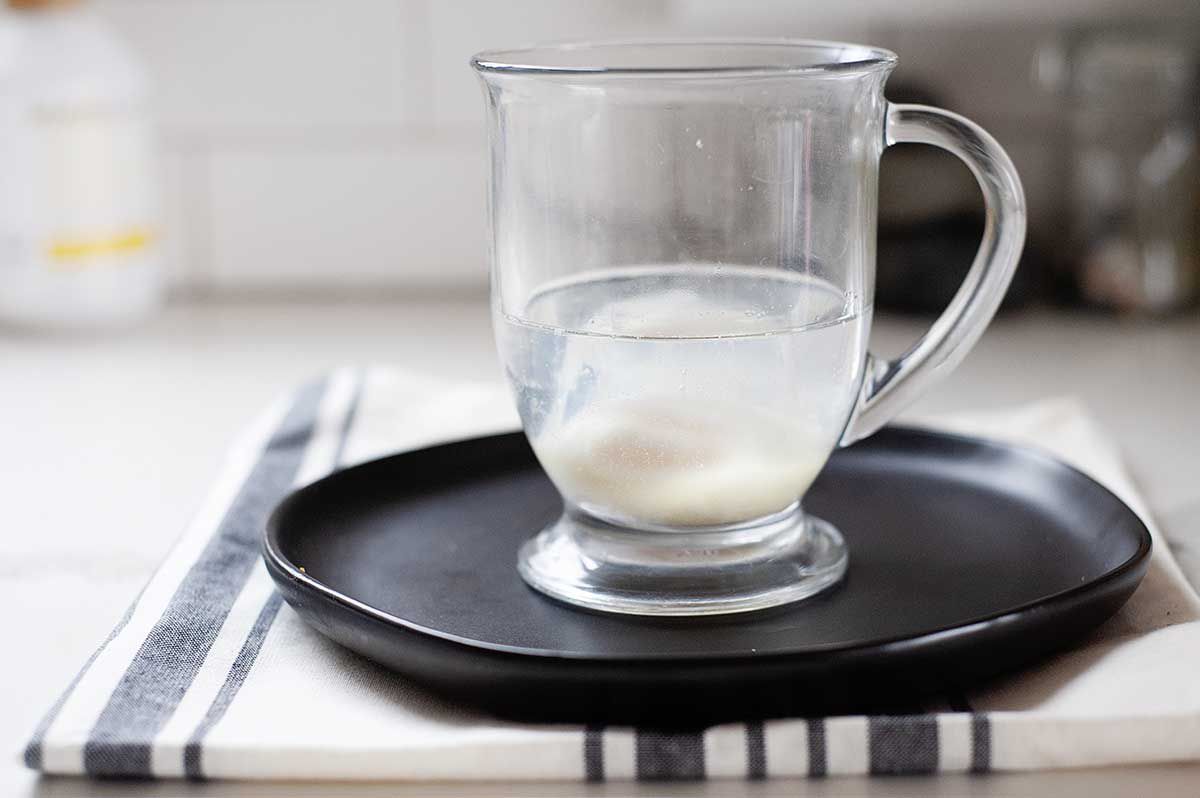 Quick poached egg and water in a glass mug. The mug is on a black plate with a linen underneath.