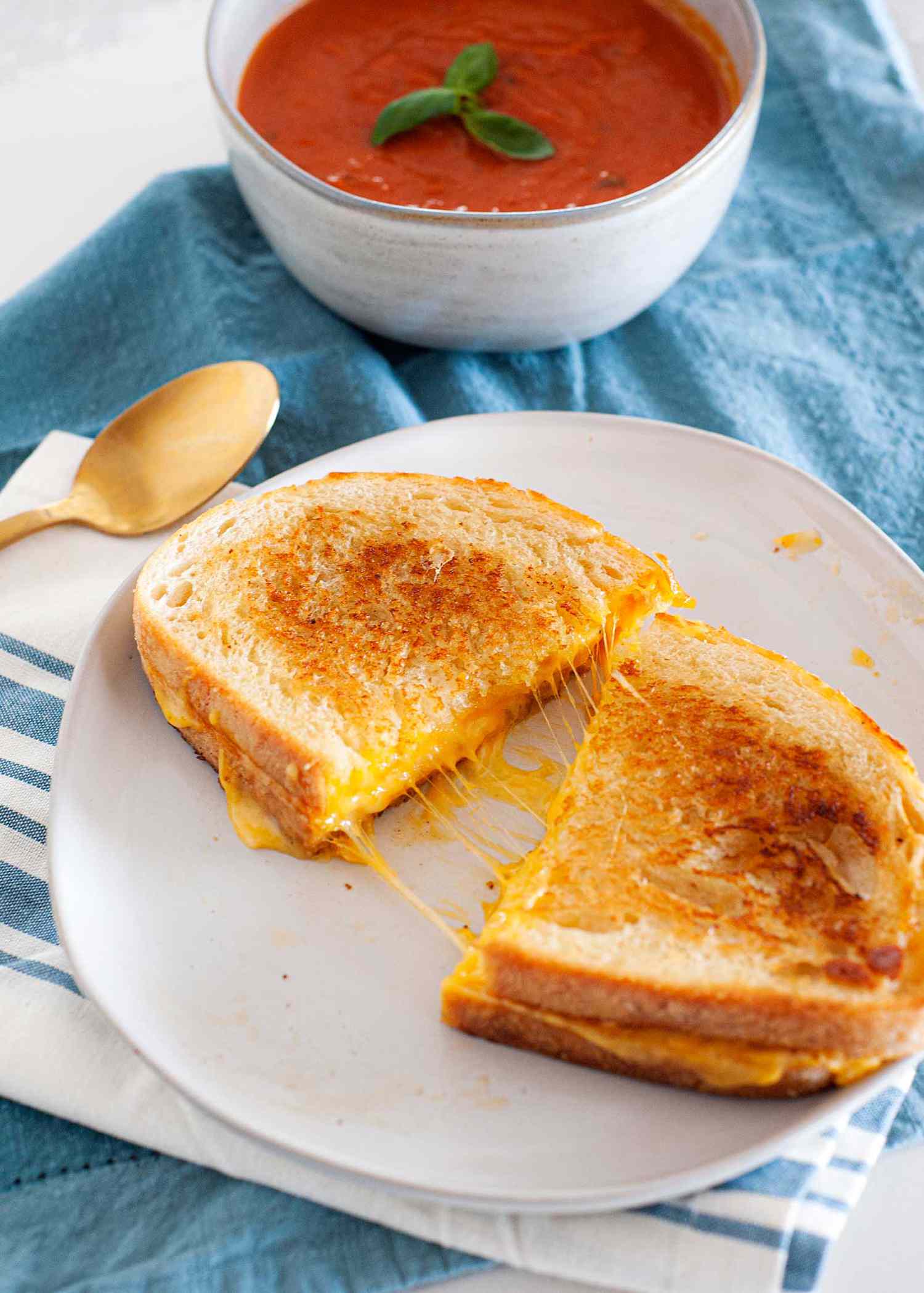 The perfect grilled cheese sandwich cut in half and split open on a plate with a blue napkin underneath it and a bowl of tomato soup in the background.