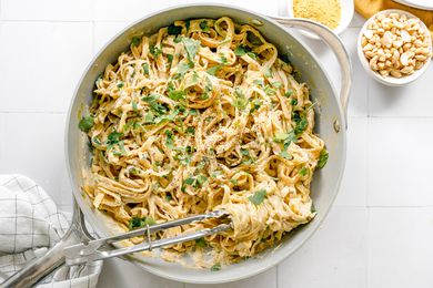 Pan of Super Creamy Vegan Pasta Topped with Parsley and Some Wrapped Around a Pair of Tongs. Next to the Pan, Two Bowls of Ingredients (One: Cashews and Two: Nutritional Yeast)