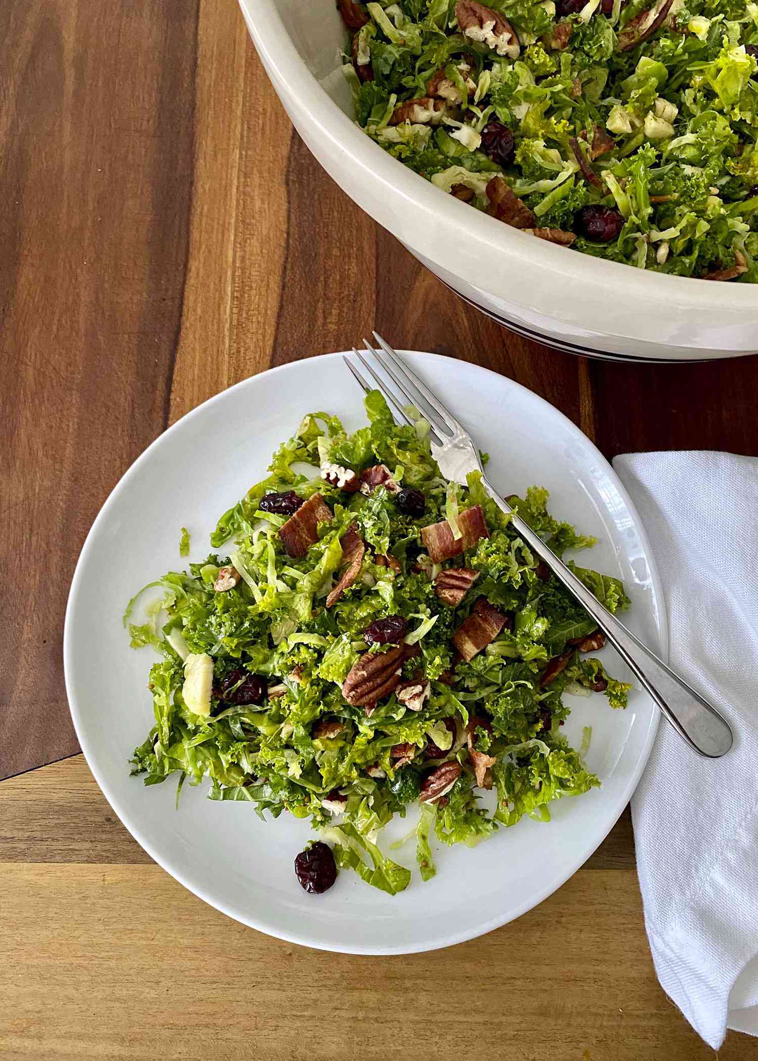 A plate and serving bowl of Raw Kale and Shaved Brussels Sprout Salad with Maple Dijon Vinaigrette.