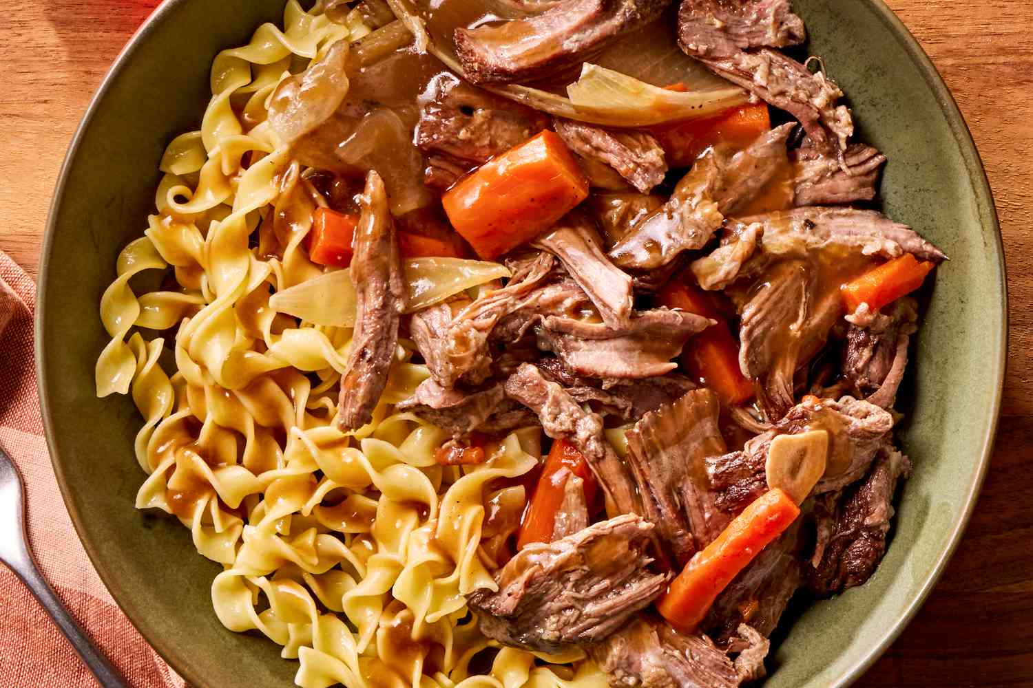Overhead view of a plate of pot roast with carrots, onions and noodles on a wooden tabletop
