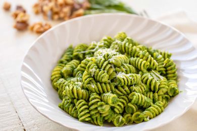 Horizontal view of a white bowl with rotini and kale pesto inside. Walnuts and basil are in the background.