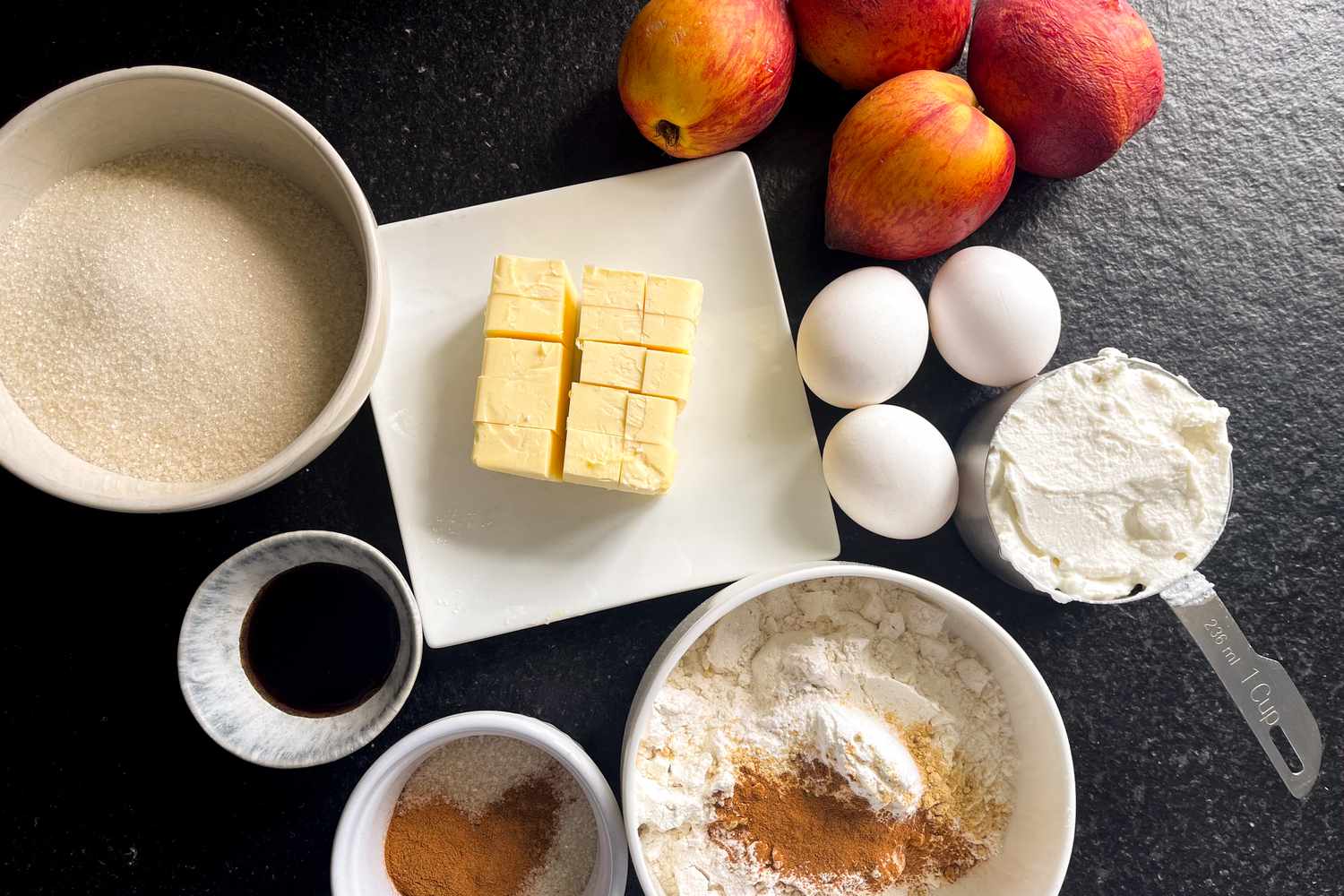 Ingredients for baking on a black surface, including butter, eggs, sugar, flour, spices, and fresh fruit