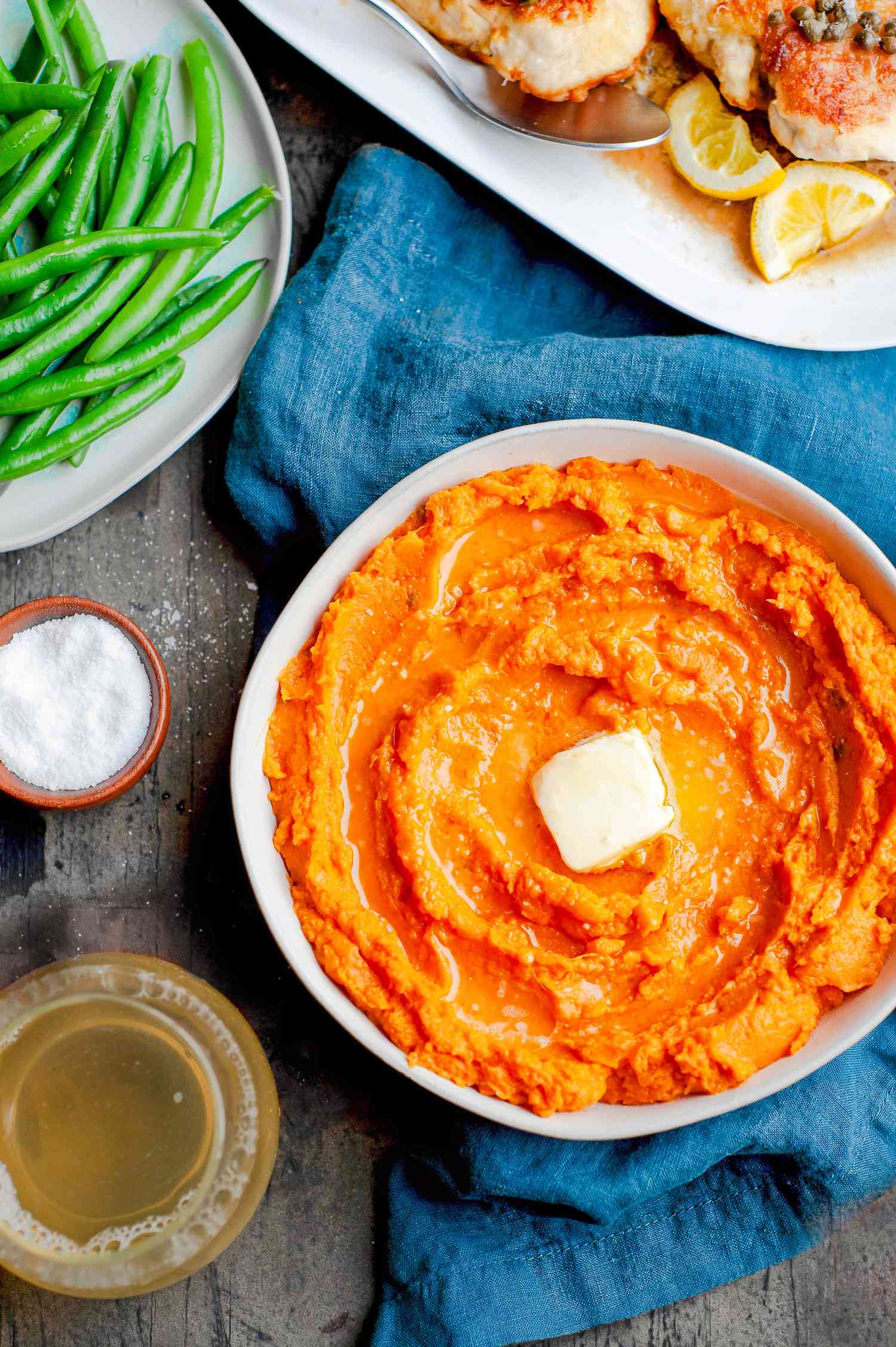Table Setting with a Bowl of Instant Pot Mashed Potatoes Topped with Butter, a Plate of Green Beans, a Platter of Chicken, a Small Bowl of Salt, a Drink, and a Table Napkin