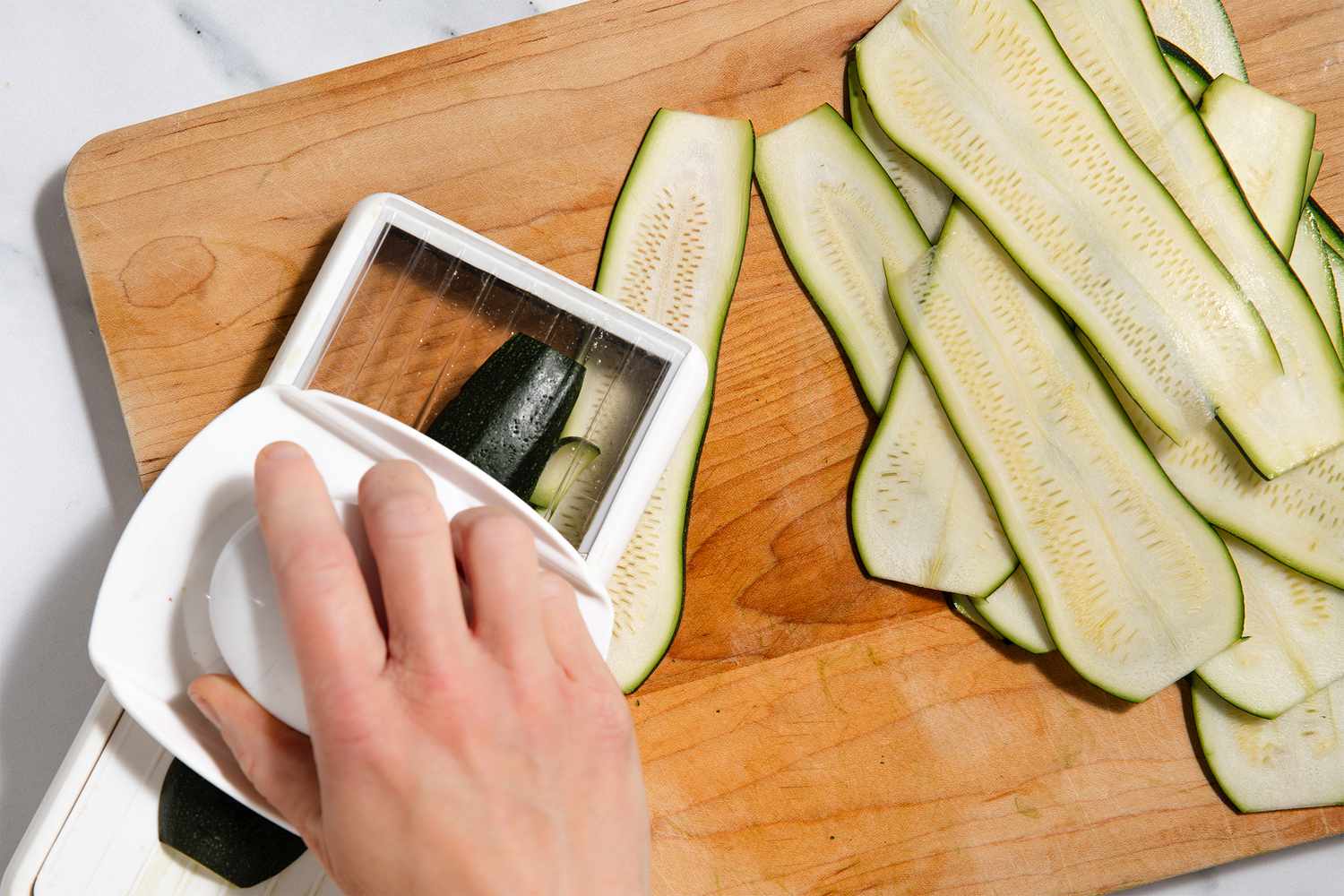 A hand slicing zucchini using a mandoline, with strips of zucchini on a cutting board