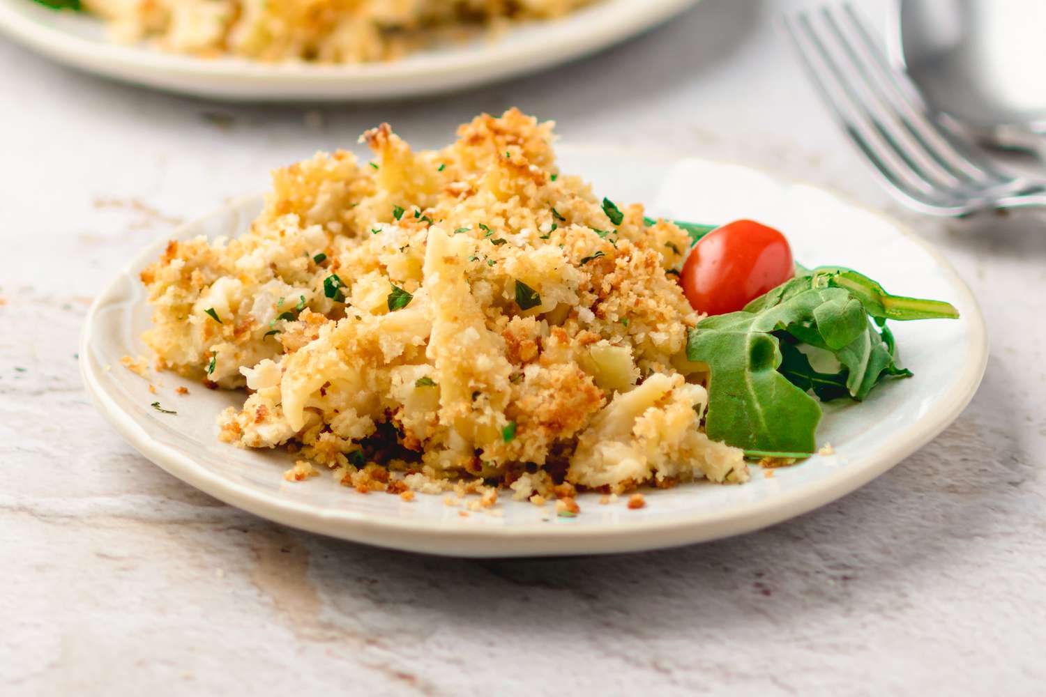 Angled view of a white plate of a serving of a casserole with topped with crushed crackers next to a cherry tomato and greens 