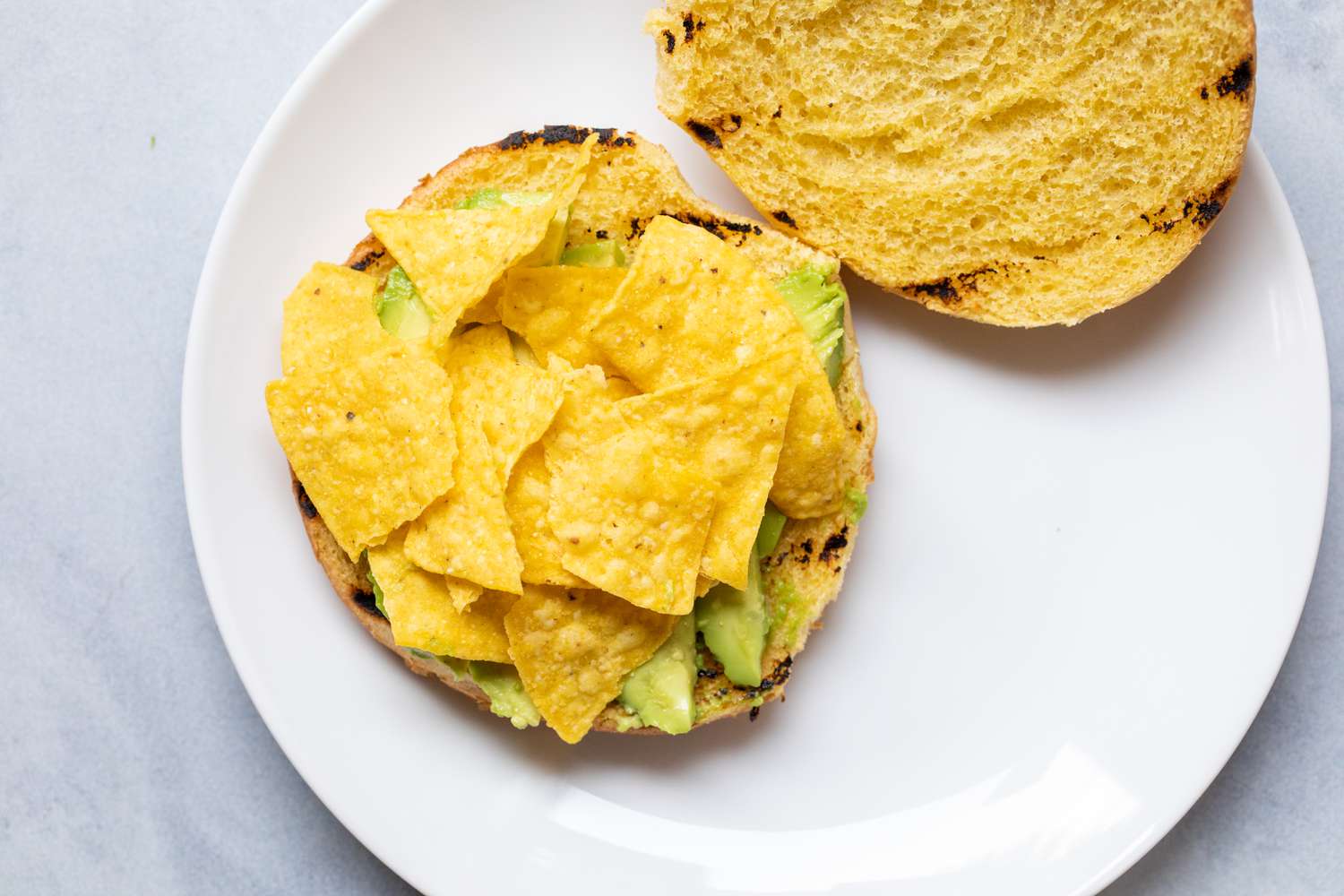 A toasted bun topped with avocado and tortilla chips on a plate to make an Avocado burger.