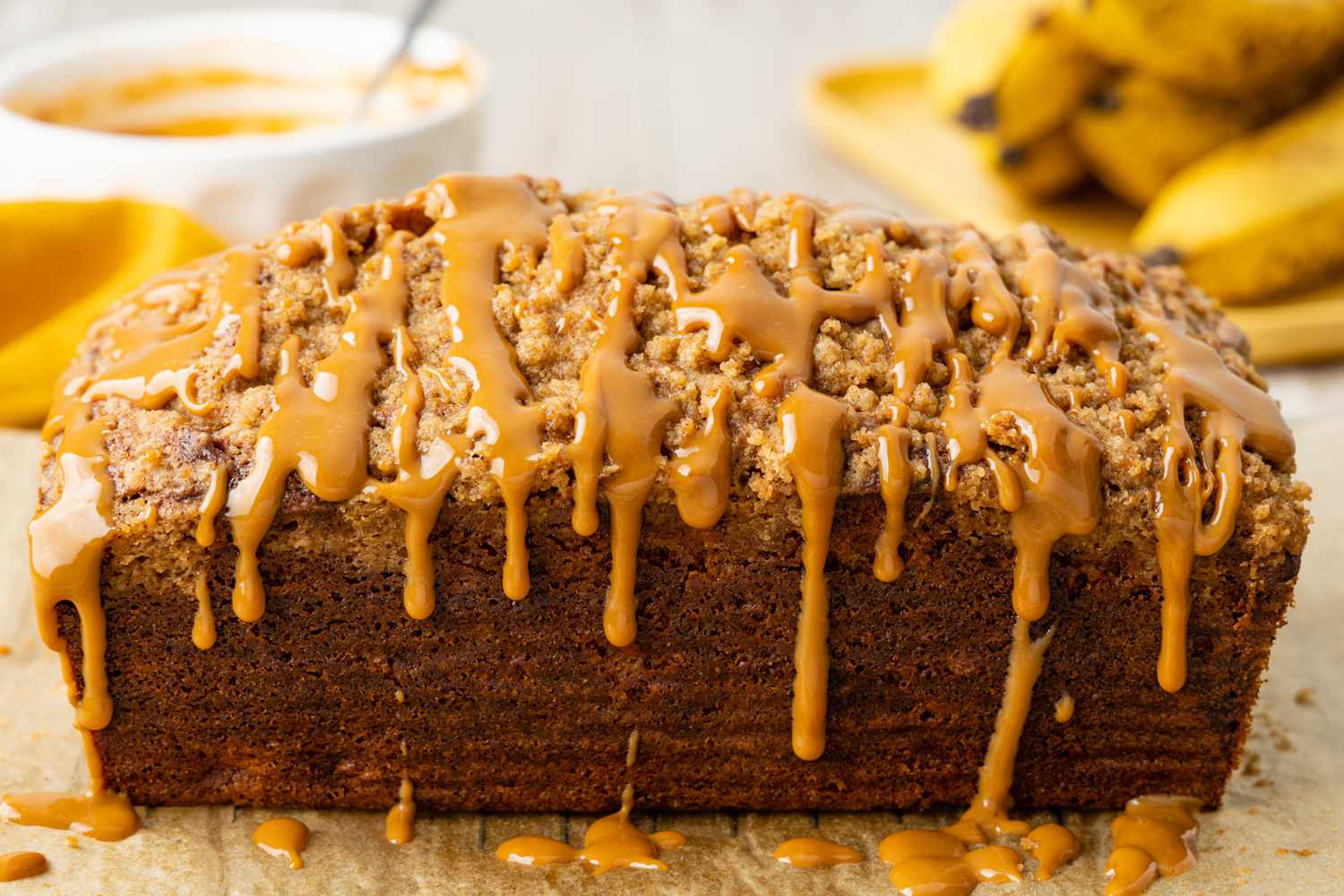 Loaf of Spiced Dulce de Leche Banana Bread With Drizzle Sitting on a Piece Parchment Paper, and in the Surroundings, a Bowl of Drizzle and Bananas
