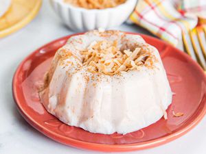 Homemade bundt shaped templeque is on a coral plate with a gold spoon set to the left. The tembleque has toasted, shredded coconut mounded in the center. Behind the plate is a striped dish towel, small white container and yellowl plate all in partial view.