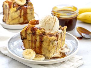 Slice of the best slow cooker banana bread pudding topped with a scoop of ice cream and drizzeld with caramel sauce on a gray plate. A jar of caramel sauce is off to the side.