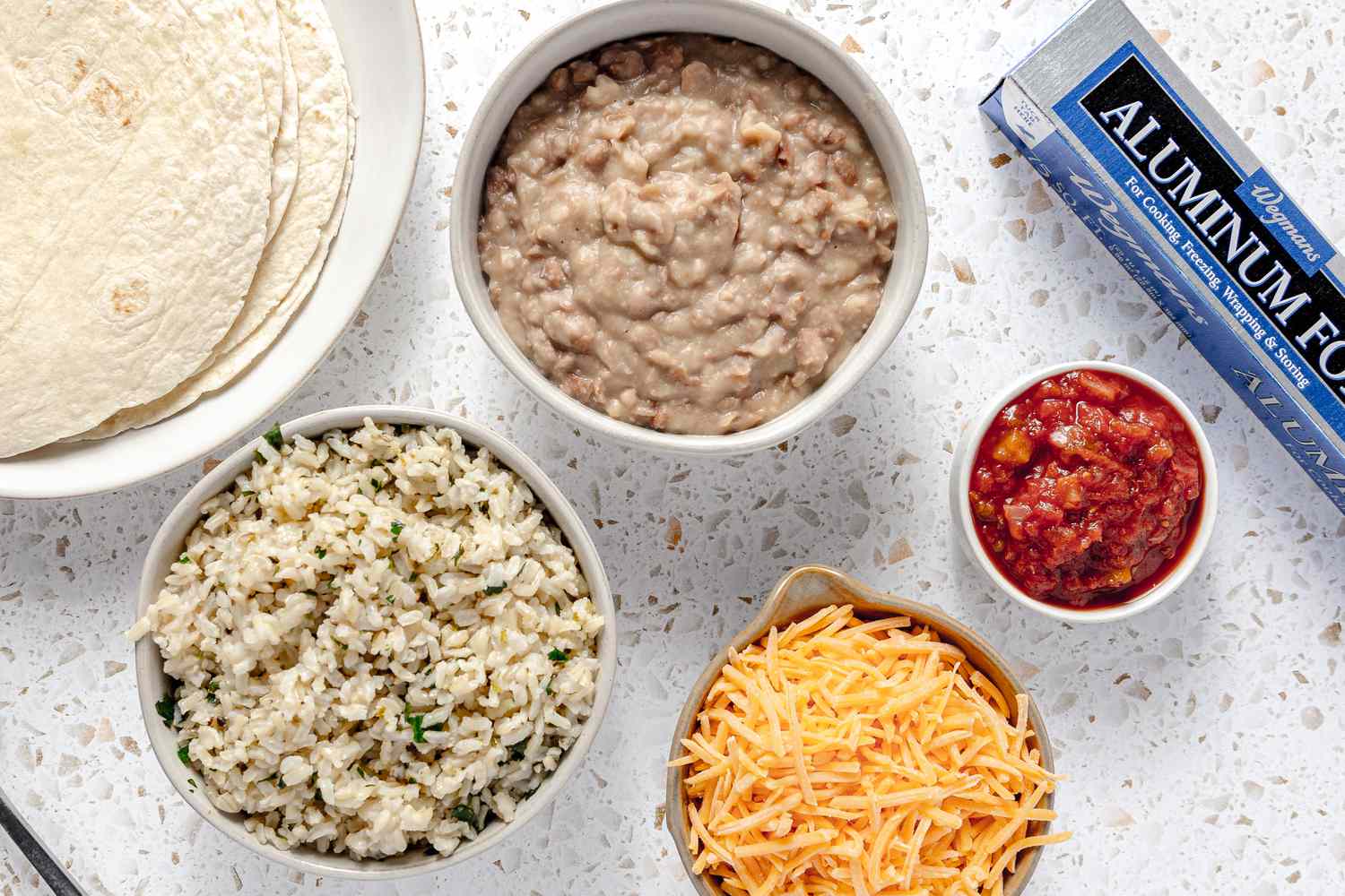 Easy Rice and Beans Freezer Burritos Ingredients on the Counter (L to R): Plate of Tortillas, Bowl of Rice, Bowl of Cooked Beans, Bowl of Cheddar, Small Bowl of Salsa, and a Roll of Aluminum Foil