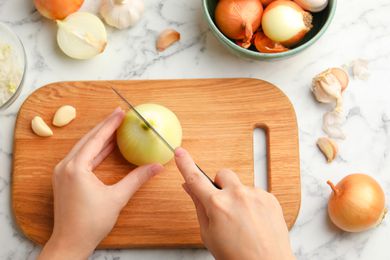 Overhead view of a woman's hands cutting an onion in half on a wooden cutting board, on a marble countertop next to other onions in a bowl and and garlic to the side
