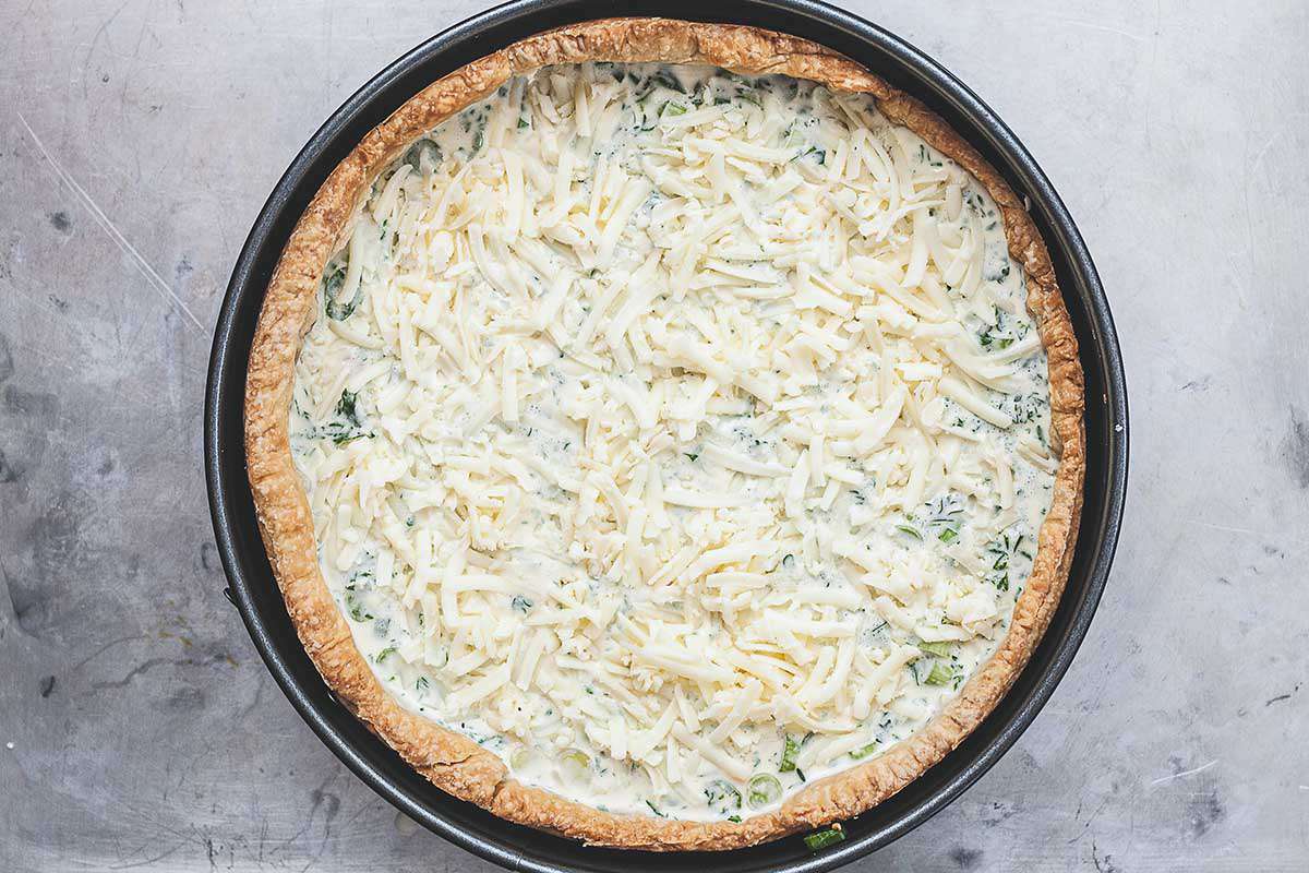 A homemade deep-dish bacon and cheddar quiche is on a countertop with an unbaked filling. The crust is golden brown and in a springform pan. Creamy filling and shredded cheese are inside the crust.
