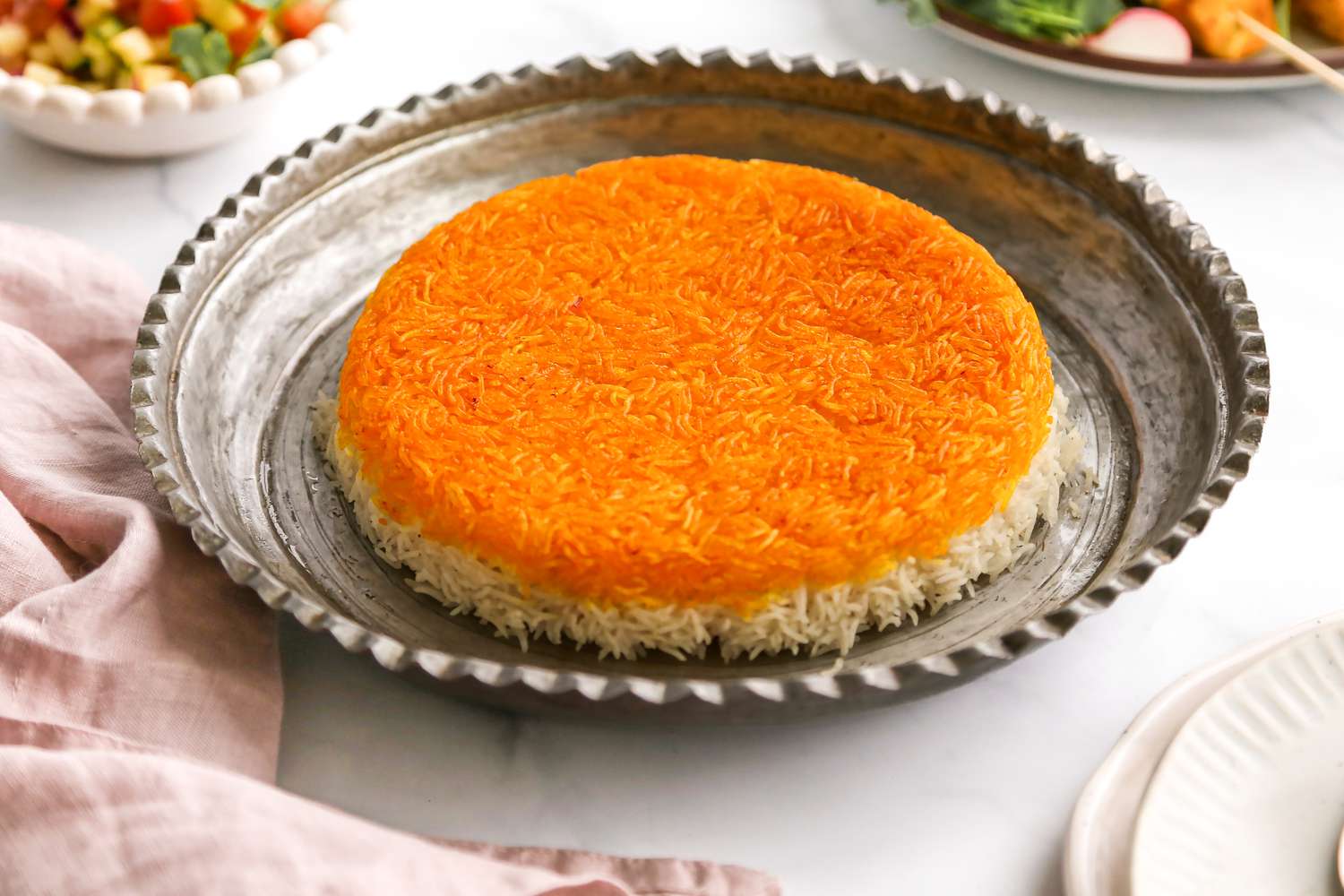 Tahdig on an Ornate Silver Platter with a Serving Spoon and Next to It, a Table Napkin, Stack of Plates, and Two Plates of Food