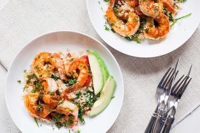 Two white plates on a white table with a stack of forks and knives to the right. Each plate has large shrimp, avocado slices, herbs and cauliflower rice inside.
