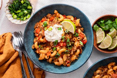 Two Bowls of One-Pot Taco Pasta Topped With Sour Cream, Sliced Scallions and a Lime Wedge, and in the Surroundings, a Bowl With Lime Wedges, a Bowl With Sliced Green Onions, a Bowl With Sour Cream, Glasses With a Drink, and a Mustard Colored Table Napkin With a Pair of Silverware