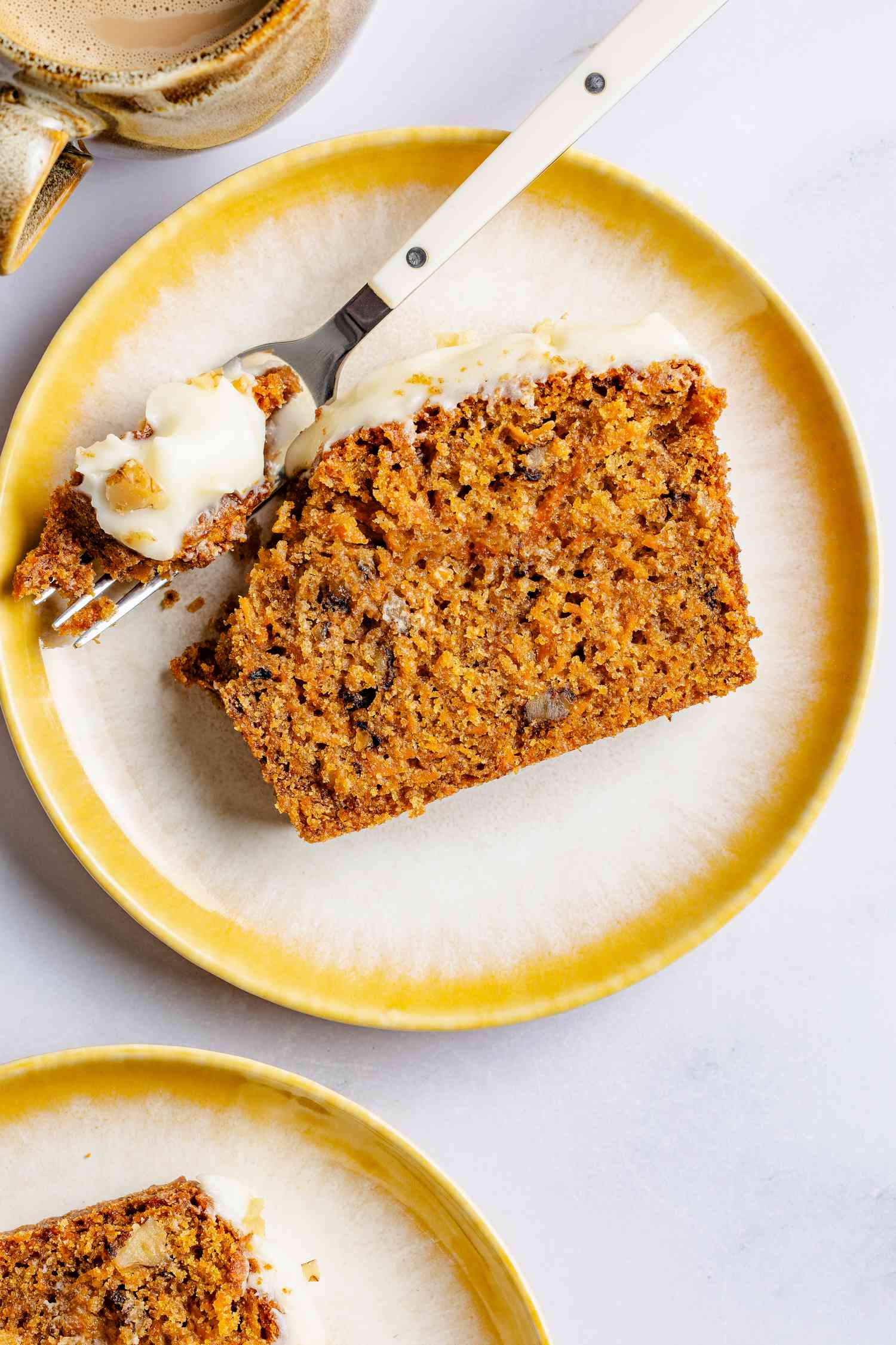 slice of carrot cake loaf on a small plate with a fork next to a cup of coffee and a plate with another slice