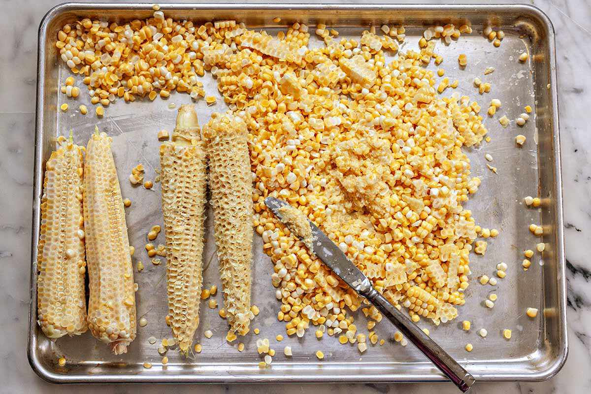 Corn kernels in a baking sheet with cobs set next to it to make a corn recipe.