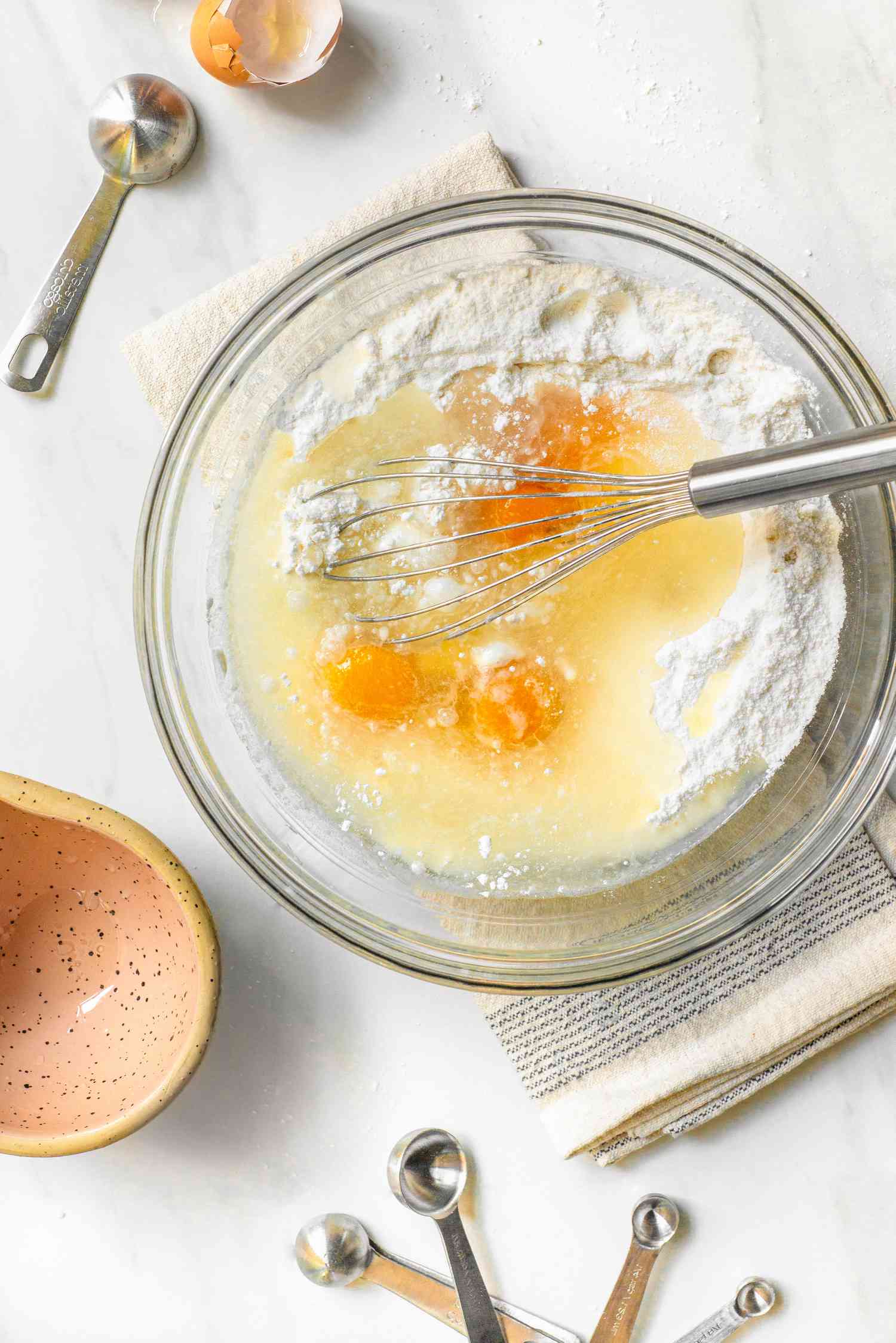 Egs and flour in a bowl with a whisk, sitting on a white kitchen towel, and in the surroundings, a measuring spoons, an egg, and a small bowl