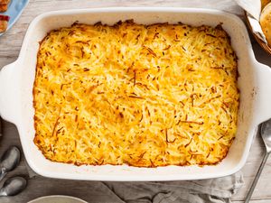Copycat Cracker Barrel hash brown casserole in a casserole dish at a table setting with a plate, a basketful of bisquits, a stack of plates, and spoons on the counter 