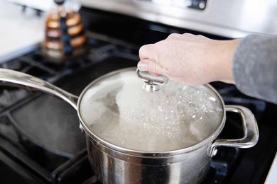 Silver saucepan with a hand grasping the lid handle. Pan sits on gas stove.