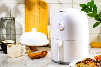 An air fryer on a kitchen counter surrounded by other kitchen tools like a set of olive wood measuring spoons, a cutting board, and two coffee mugs
