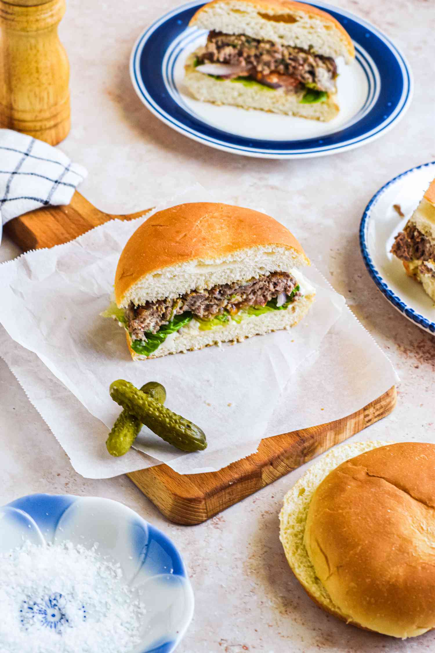 Halved Blue Cheese Burger on Parchment Paper with Pickles Surrounded by Plates with More Halved Sandwiches and a Small Dish with Flaky Salt