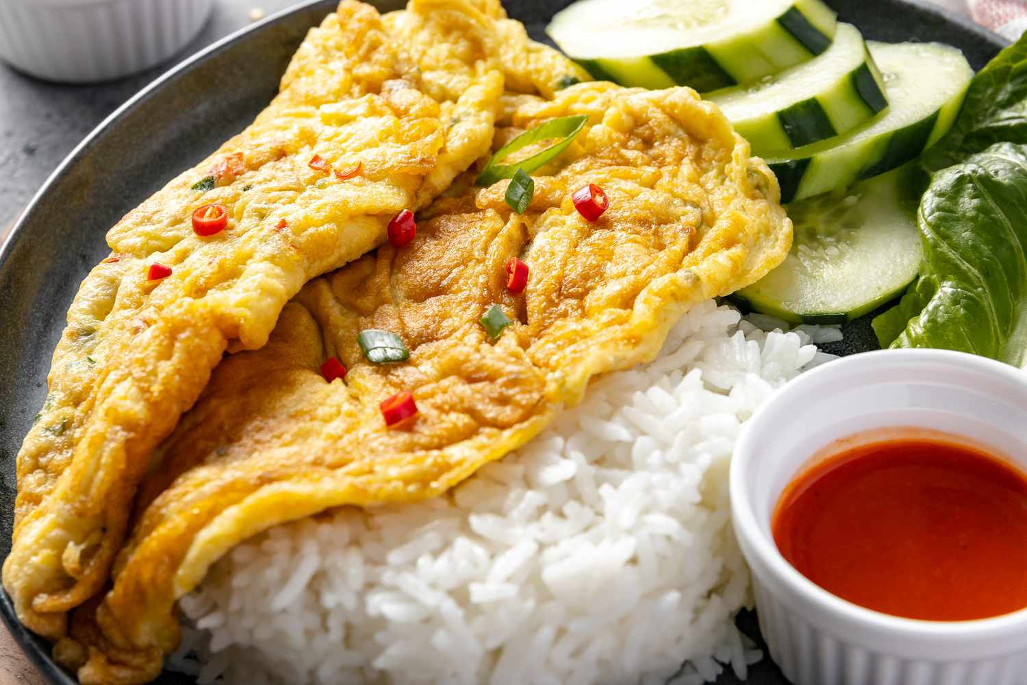 Close Up: Plate of Kai Jeow (Thai Omelet) with Cucumber Slices, Romaine Leaves, Rice, and a Small Bowl of Sriracha