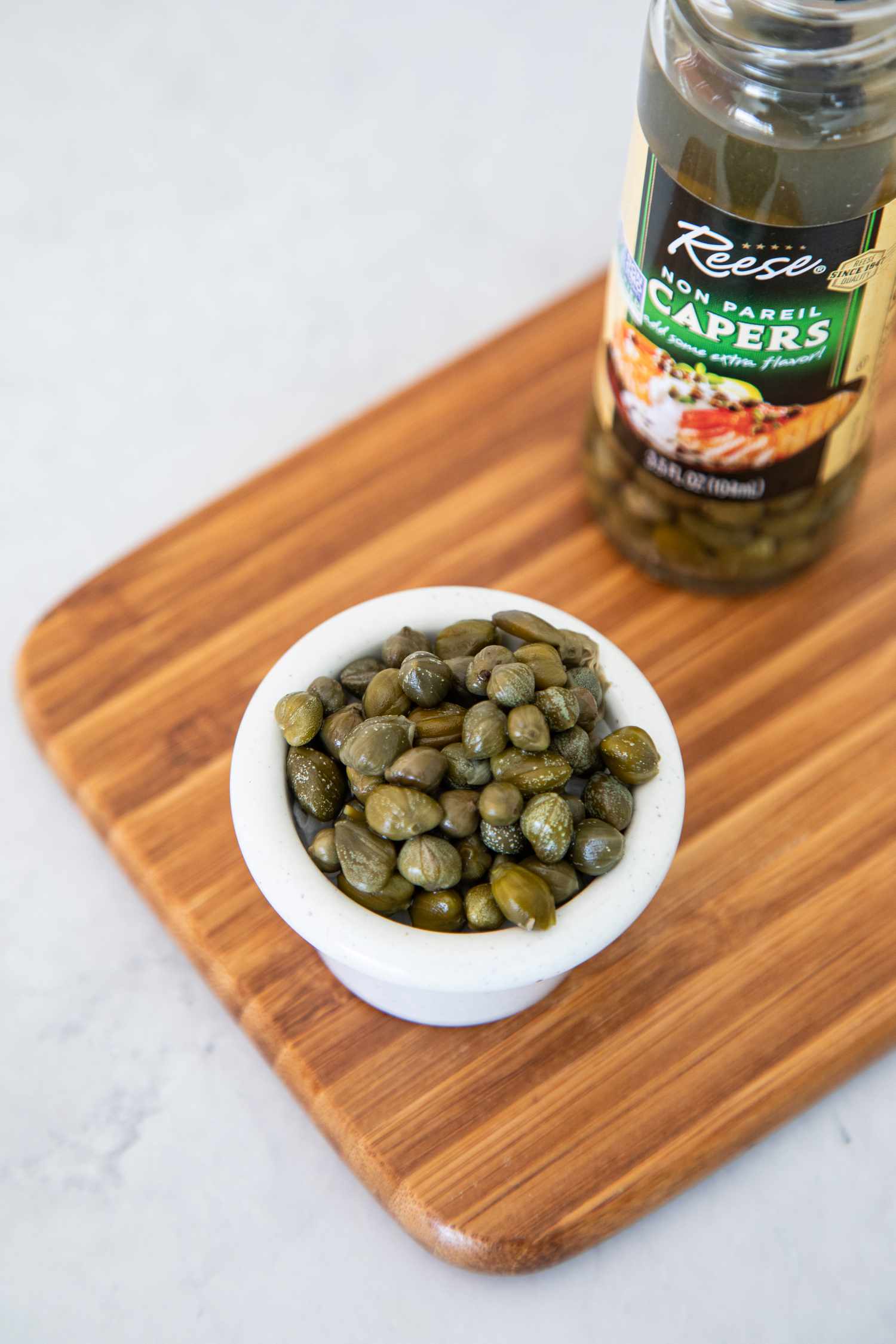 Small Bowl of Capers with the Jar Behind It, and Both Are on a Wooden Board