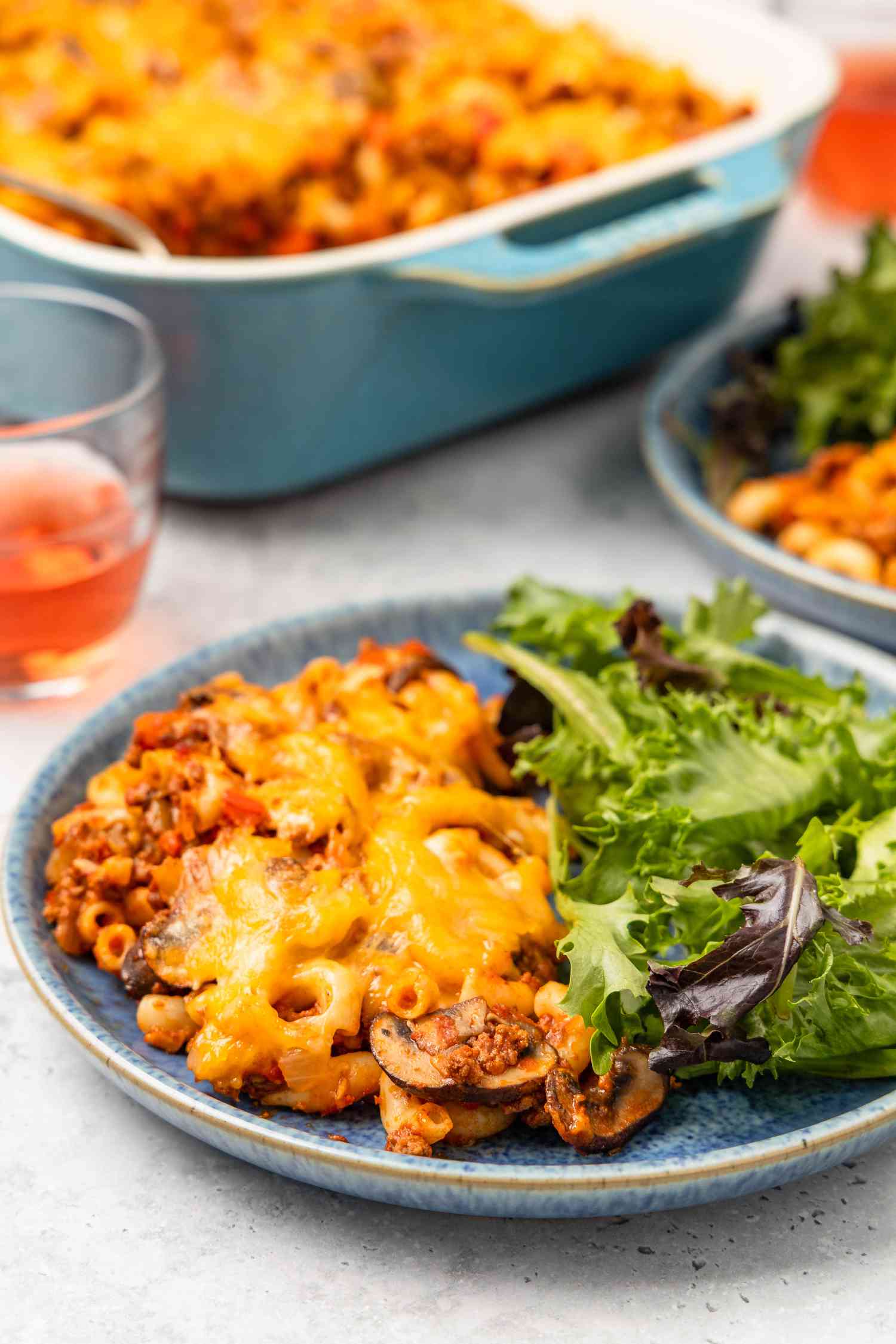 Serving of Johnny Marzetti casserole and mixed greens on a plate at a table setting with more casserole in a casserole dish, another plate with another serving and a drink 