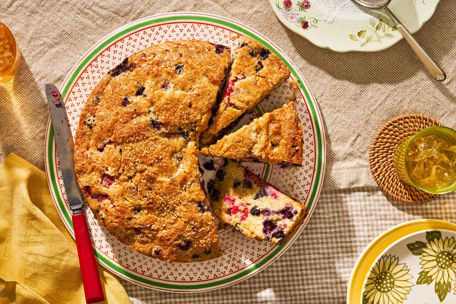berry buckle (some of cut into slices) on a plate next to a stack of plates, a plate with a powdered sugar duster, and a glass, all on a cozy table setting (cottagecore vibes)
