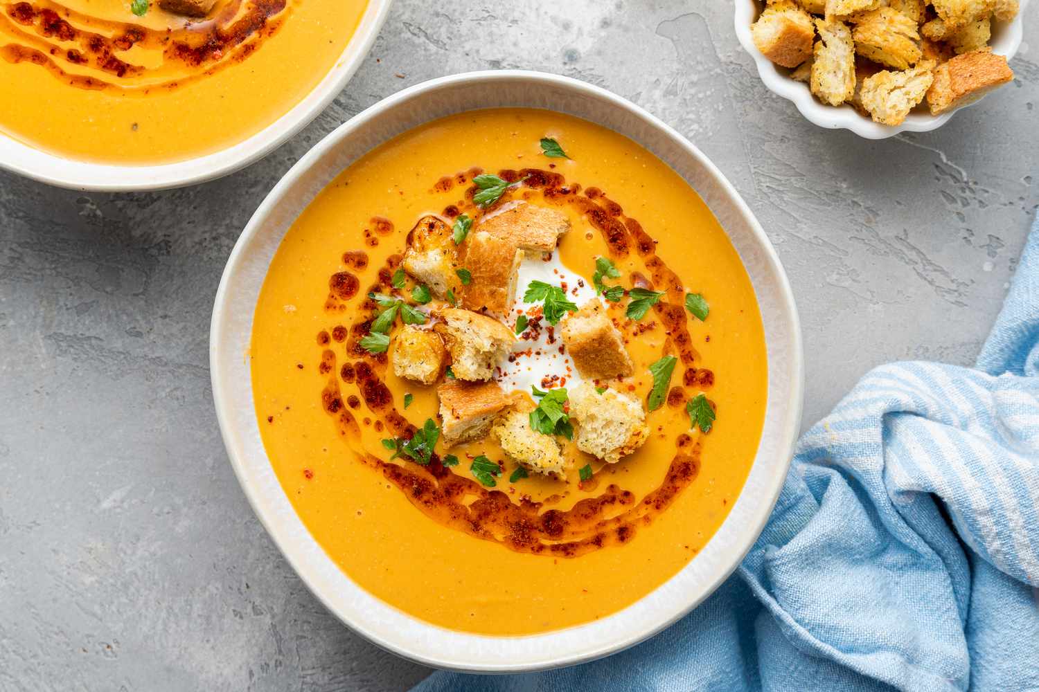 Bowls of Mercimek Corbasi (Turkish Lentil Soup) Topped with Greek Yogurt, Croutons, Pul Bibir Butter, and Parsley, and Next to It, a Kitchen Towel and a Small Bowl of Croutons