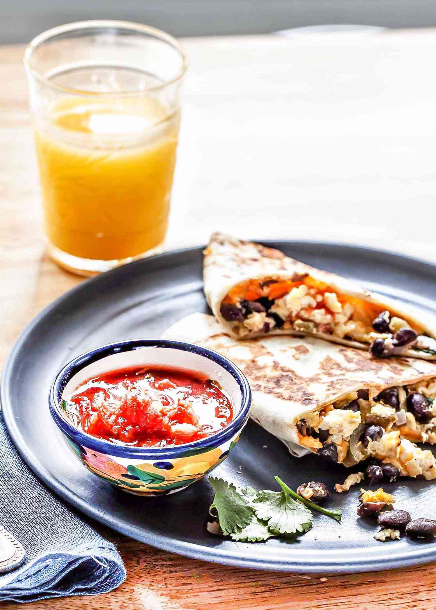 A blue plate with two quick and easy breakfast quesadillas with the scrambled egg, black bean and cilantro filling tumbling out the side. A small bowl of salsa is on the left of the plate and a glass of orange juice is above the plate on the left.