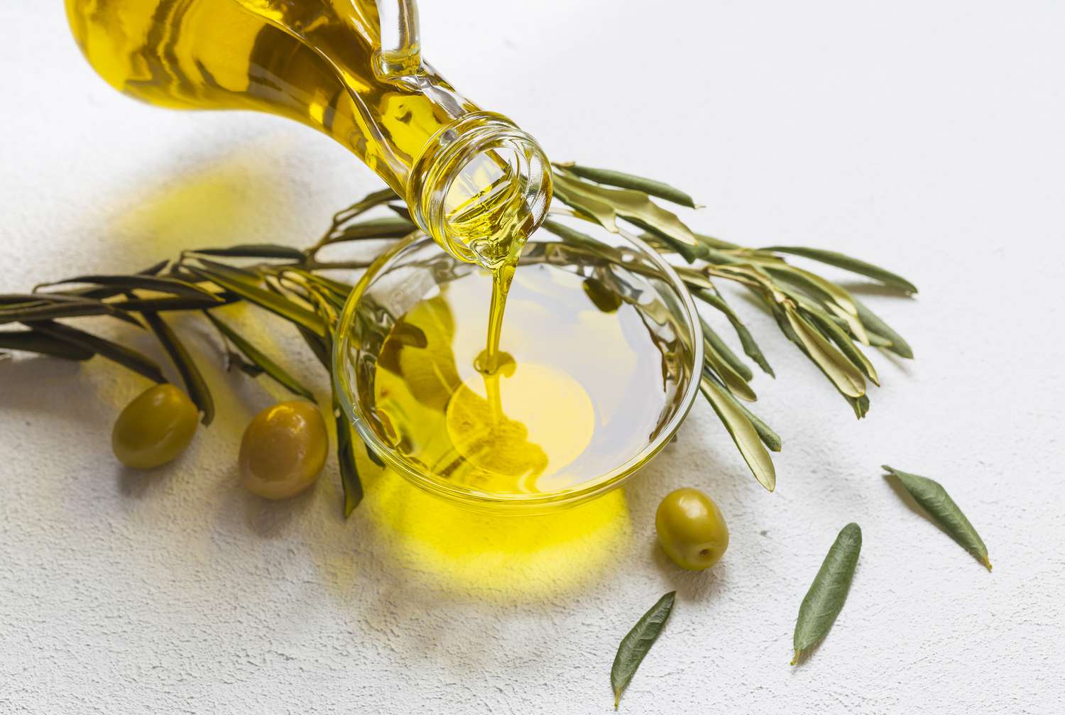 Olive oil being poured from a glass cruet into a small bowl. A sprig of olive leaves and a few green olives sit to the side.