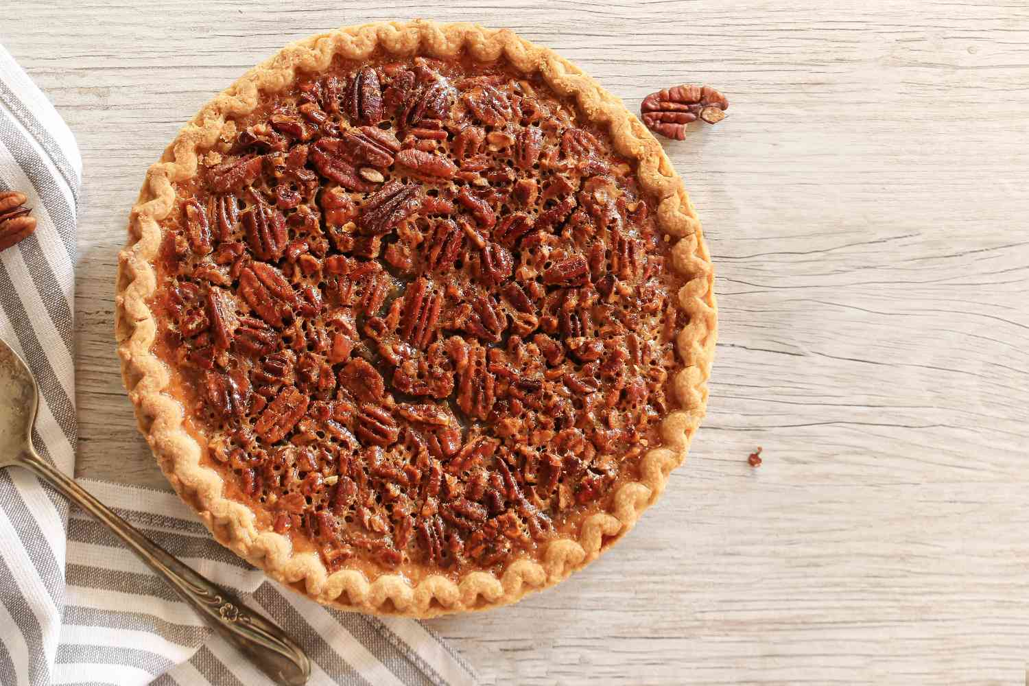 Overhead view of a pecan pie next to a striped towel and pie server all on a wooden tabletop