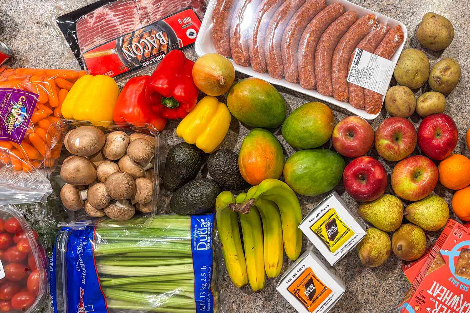 A grocery haul on a kitchen counter showing sausages, bacon, bell peppers, bananas, mangoes, potatoes, apples, pears, avocados, mushrooms, carrots, celery, and plum tomatoes