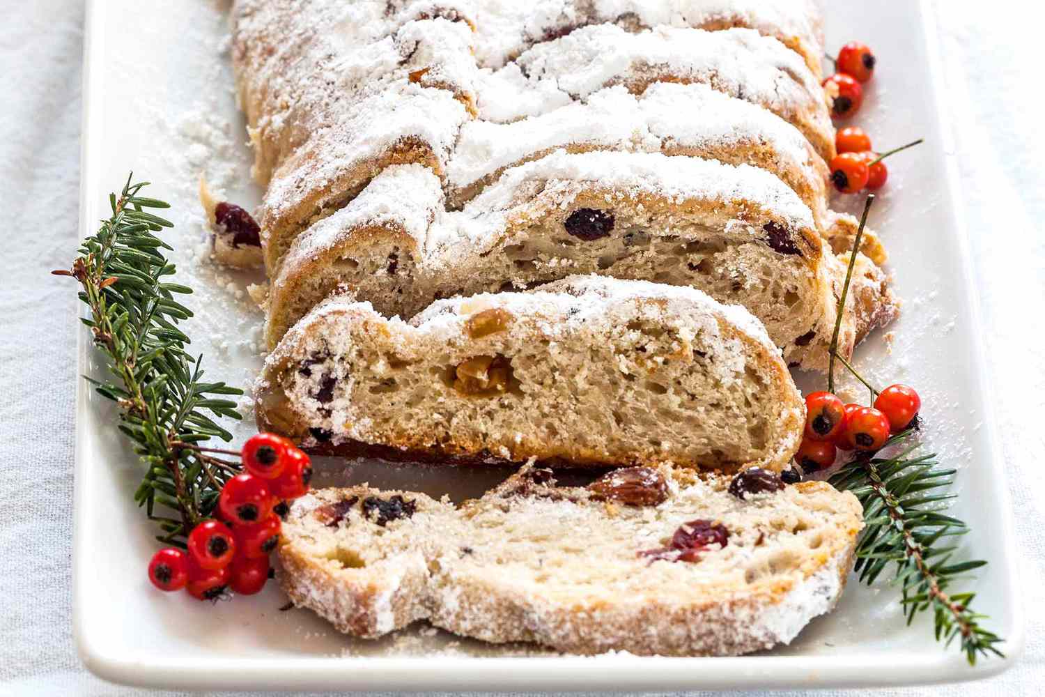 Stollen Christmas Bread filled with rum-soaked dried fruit coated with powdered sugar and sliced on a platter with a fine mesh sieve with powdered sugar with pine sprigs and berries nearby.