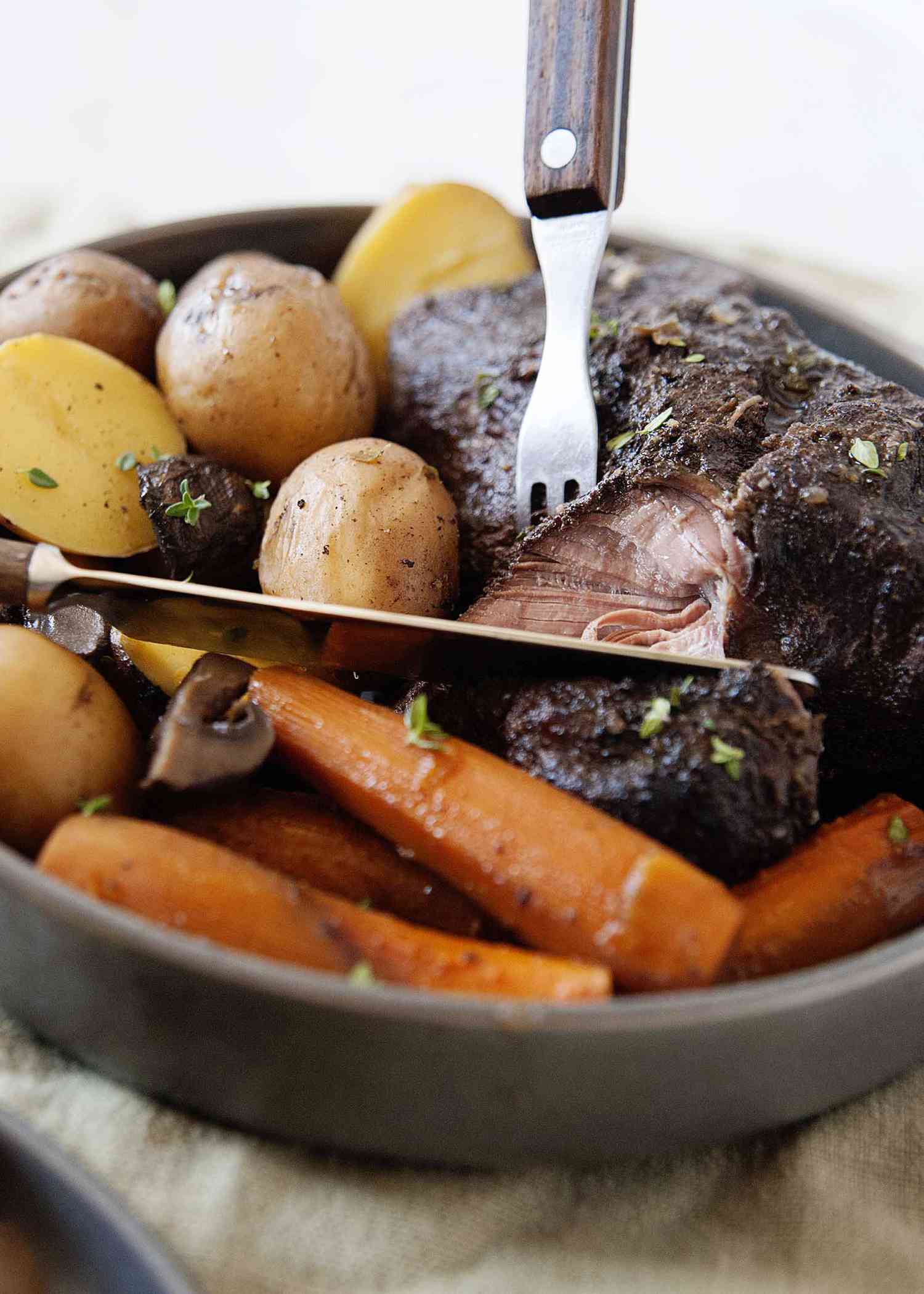 Close up of tender crock pot roast made in a slow cooker with gracy, cooked carrots and potatoes with a fork and knife.