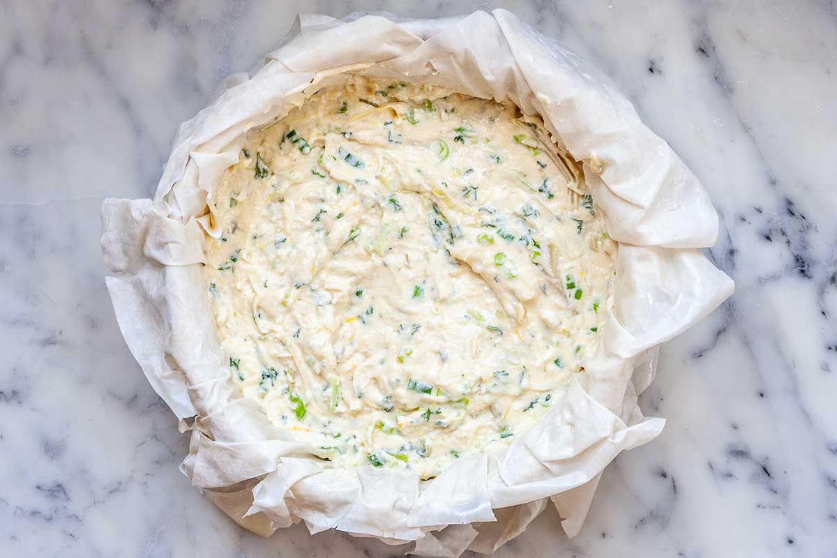 A cake pan is on a marble countertop. Unbaked phyllo dough is layered underneath a creamy cheesy artichoke pie filling. The phyllo is draped over the sides of the pan. Green flecks are visible in the pie filling.