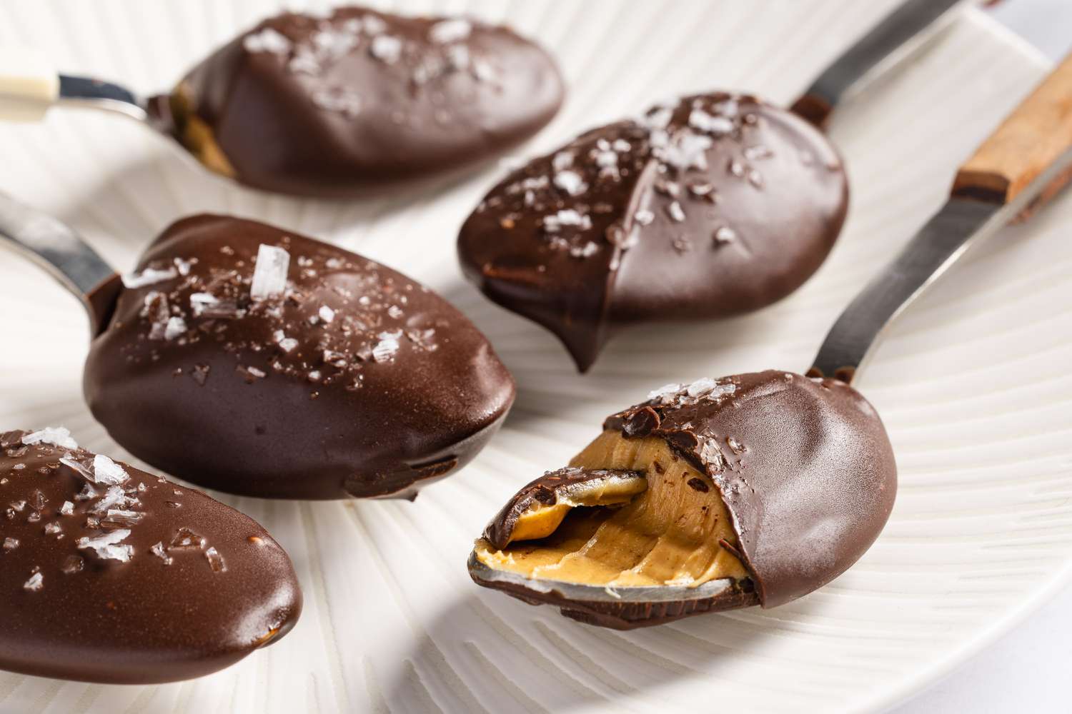 Side view closeup of chocolate and sea salt covered peanut butter on spoons, with a bite taken of one to show the creamy peanut butter inside