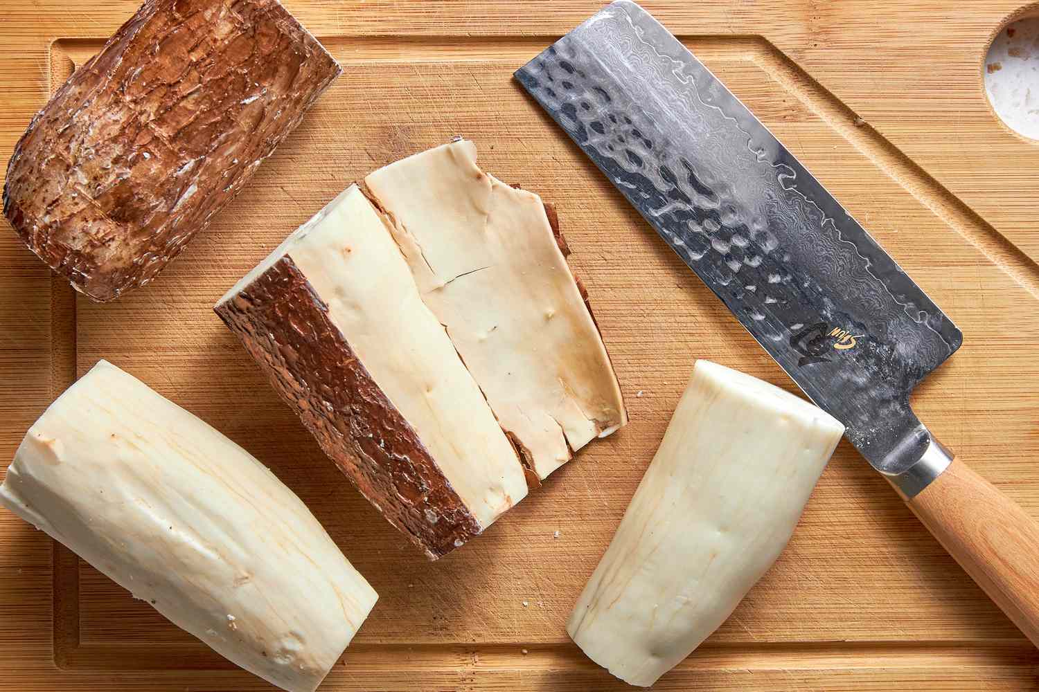 Yuca / Cassava Root With the Outer Skin Slightly Removed Next to More Yuca Roots (Some With Skin Removed and Some With Skin Intact) and a Knife, All on a Cutting Board