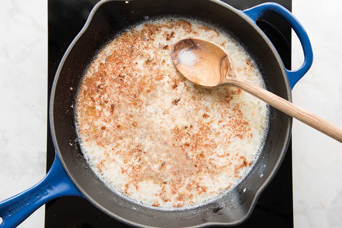 A black cast iron skillet will spices, milk and cheese being stirred together with a wooden spoon for gameday queso