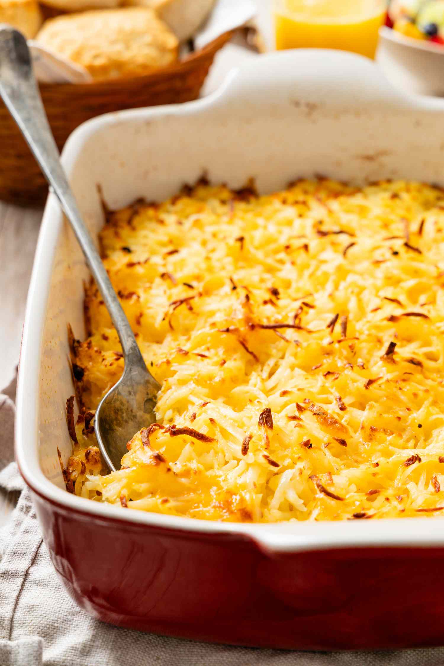 Spoon digging into copycat Cracker Barrel hash brown casserole in a casserole dish with a basketful of biscuits and a glass of orange juice in the background