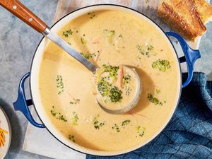 Pot of copycat Panera broccoli cheddar soup with a ladle raising some of it, and in the surroundings, a bowl of shredded cheese, a piece of baguette, and a blue kitchen towel