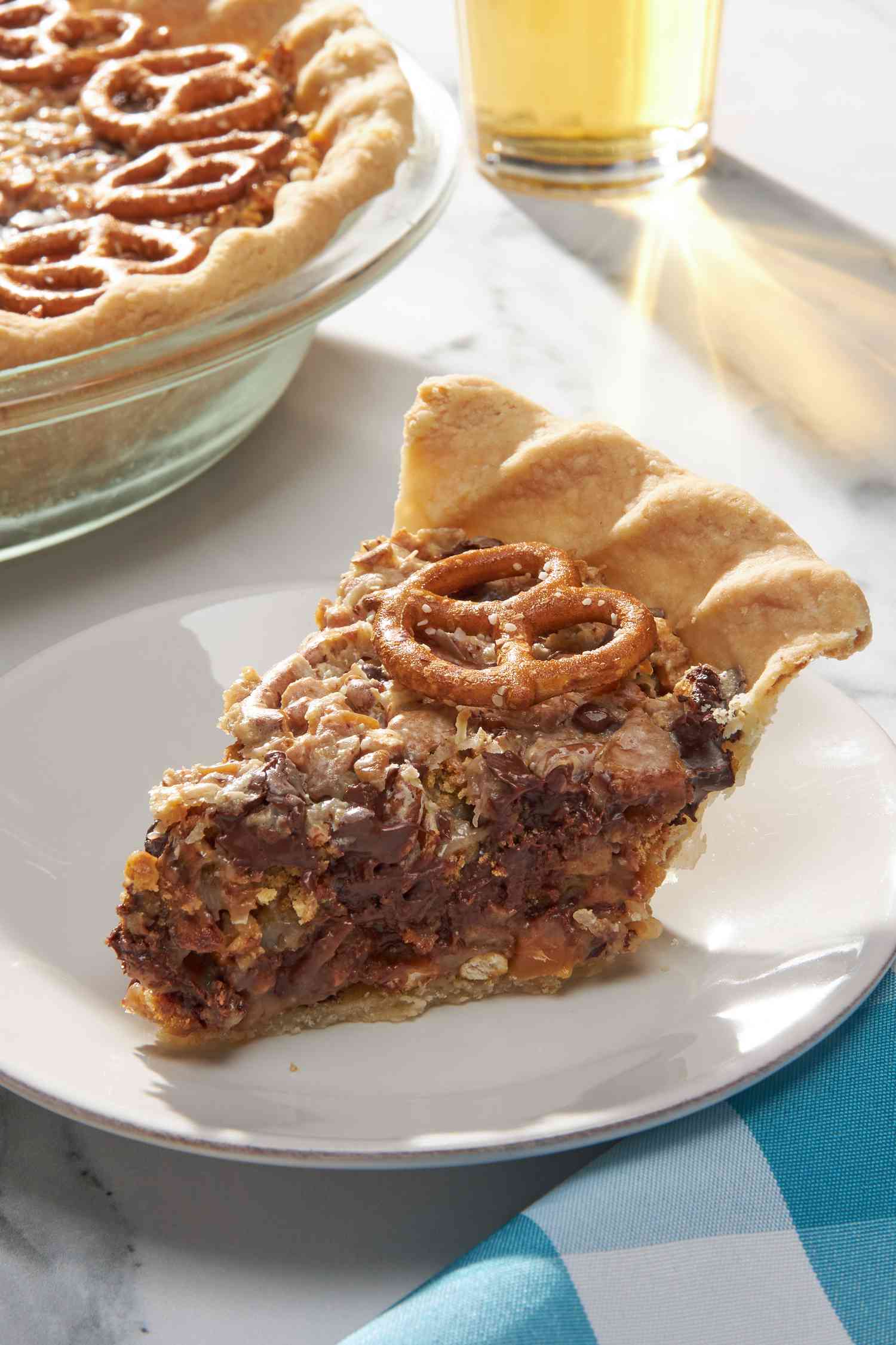 Slice of Texas trash pie on a small plate with a fork, and in the surroundings, a pie dish with more pie, a glass of beer, and a kitchen towel